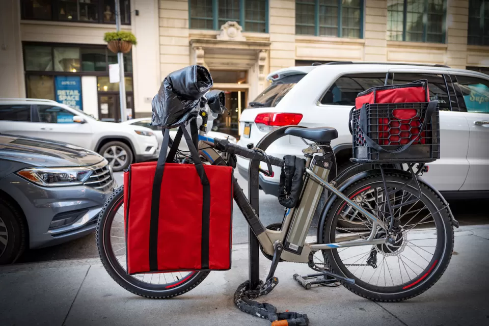 Black e-bike with red food delivery bags locked at bike rack on New York City street.